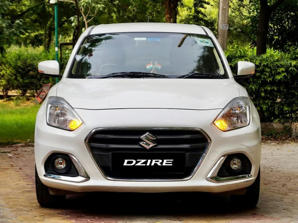 Suzuki Dzire blanche vue de face, feux de jour allumés, garée devant un parc arboré
